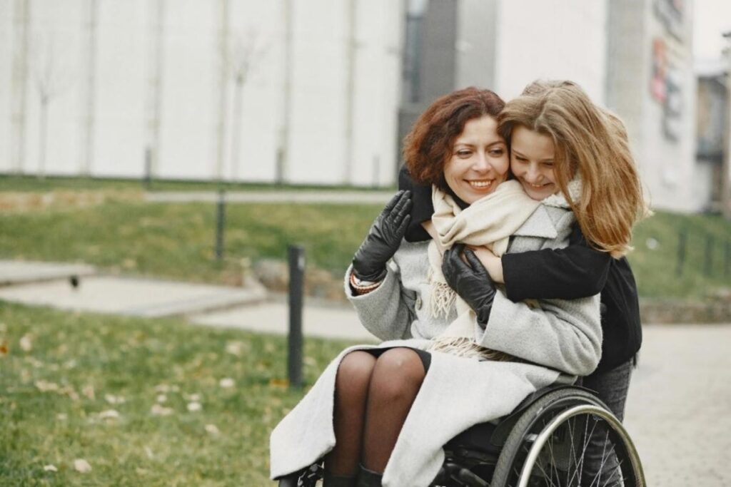 [Image description: A mother with auburn hair seated in a wheelchair while hugging her smiling, young daughter on a bright fall day.
Attribution: Gustavo Fring, Pexels (December 2020).]