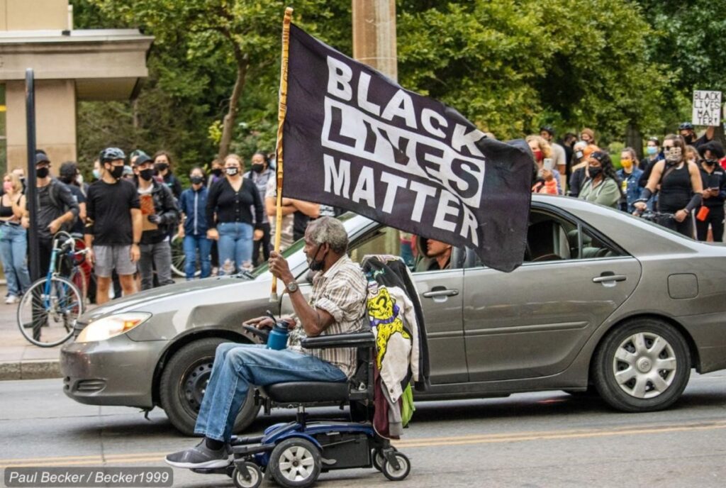 [Image description: A gray-haired Black man rolls down the street in a power chair while flying a “Black Lives Matter” flag at a protest against police brutality and racism.
Attribution: Paul Becker (2020).]