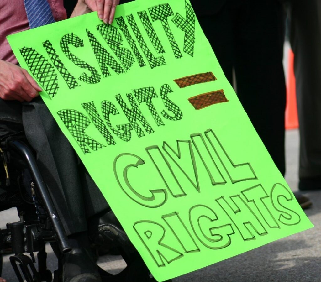 [Image description: A wheelchair user holding a protest sign that reads “Disability Rights = Civil Rights.”
Attribution: The Leadership Conference on Civil and Human Rights (2008).]
