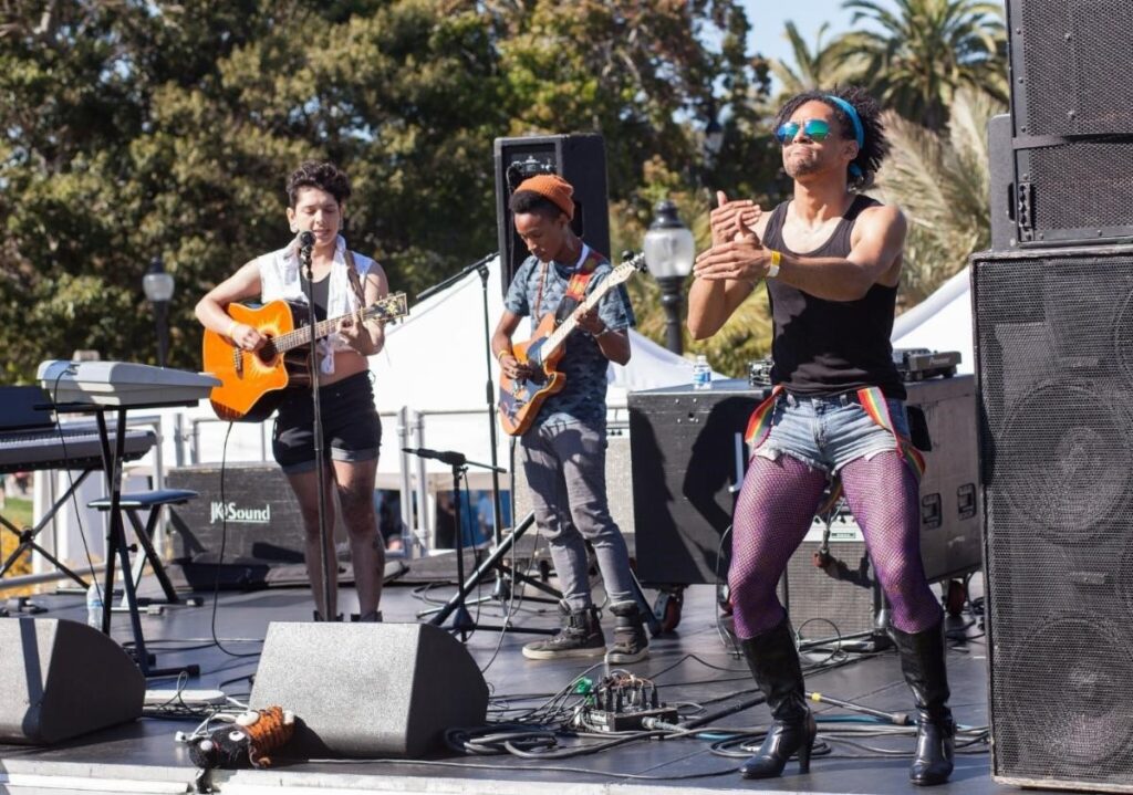 [Image description: The musical group Caveta Envinada and a sign language interpreter perform on stage at the San Francisco Trans March 2016.
Attribution: Pax Ahimsa Gethen via Wikimedia Commons (2016).]