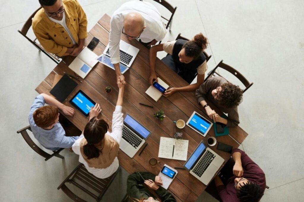 [Image description: Overhead view of a diverse group of eight coworkers sitting around a wooden table in a business meeting, with each person using different tools and technologies to meet their needs. Two coworkers are shaking hands.
Attribution: fauxels, Pexels (2019).]