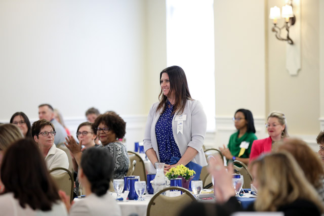 Woman stands in midst of seated people at tables.