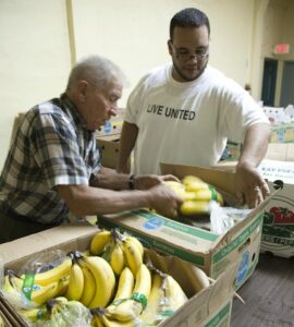 Man in Live United t-shirt helps older man select bananas.