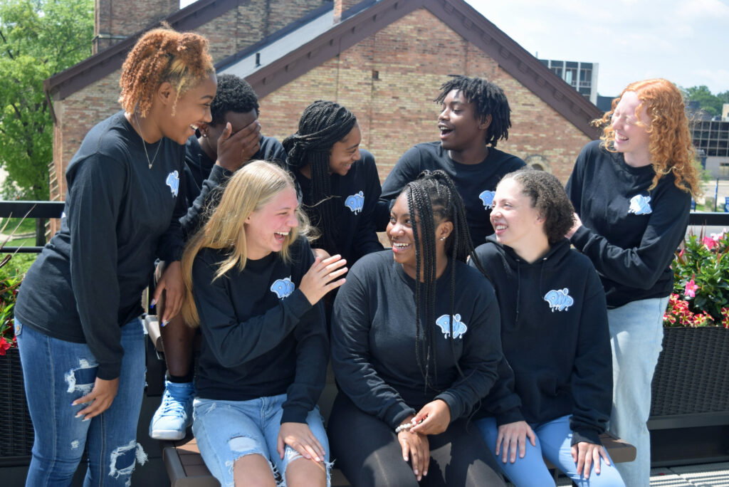 Eight teens sit on a bench and laugh while posing for photos