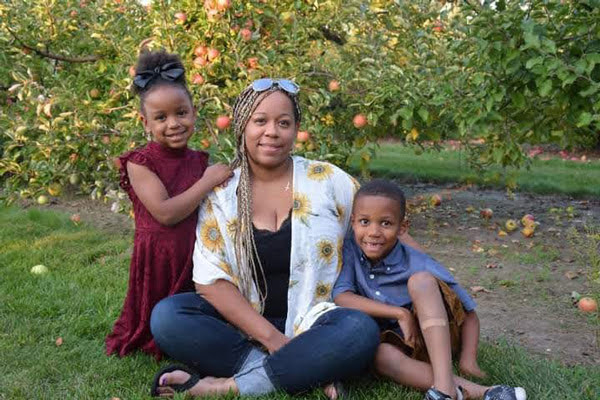 A mom and her two kids pose sitting in the grass in an apple orchard.