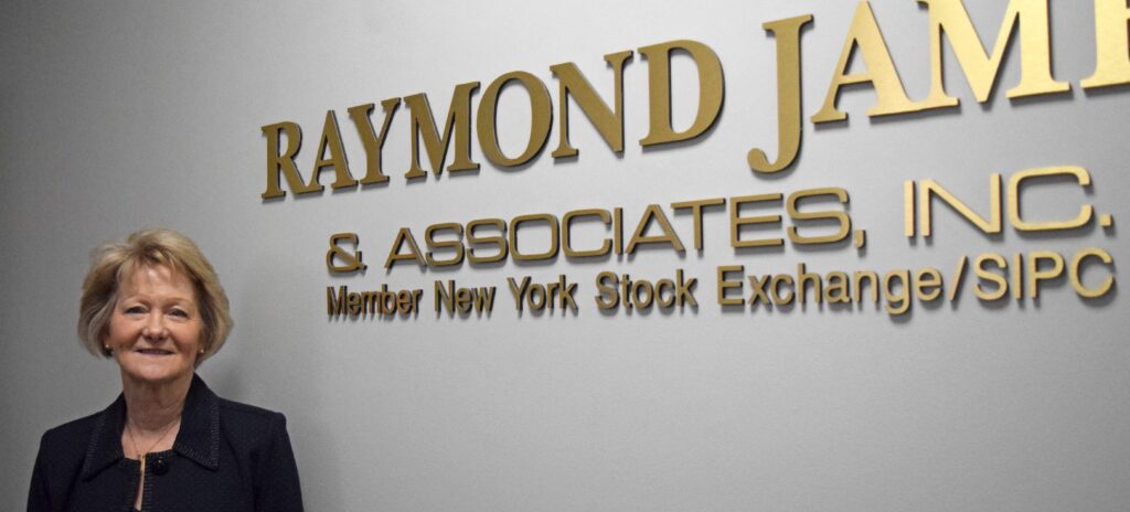Woman in black suit stands beside lettering on a wall that says Raymond James