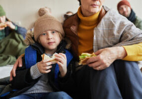 Close up of Caucasian little boy with family in refugee shelter eating sandwich and looking at camera