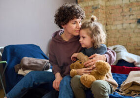 Young woman giving hug to her cute little son with brown soft teddybear while both sitting on sleeping place prepared for refugees